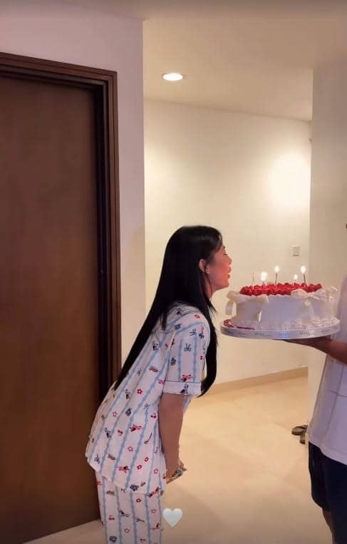 Seorang wanita berambut panjang mengenakan piyama sedang meniup lilin di atas kue ulang tahun dengan hiasan stroberi di dalam ruangan.