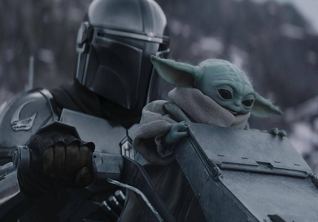 Karakter Mandalorian mengenakan baju zirah perak mengendarai speeder bersama Grogu di latar pegunungan bersalju dalam cuplikan film.