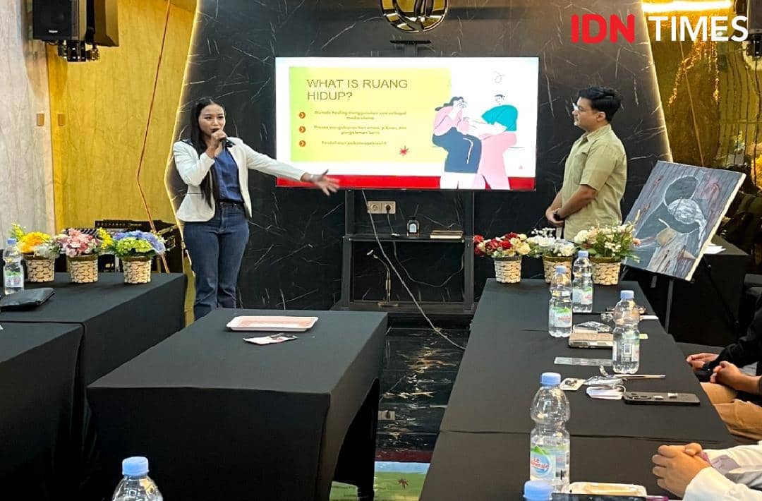 Seorang perempuan mempresentasikan topik berjudul 'What is Ruang Hidup?' di depan layar, didampingi seorang pria di ruangan dengan meja dan bunga hias.