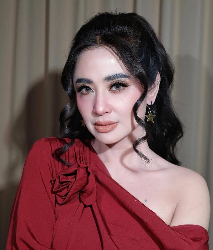 Seorang wanita berambut hitam panjang bergelombang mengenakan gaun merah dengan hiasan bunga di bahu dan anting berbentuk bintang.