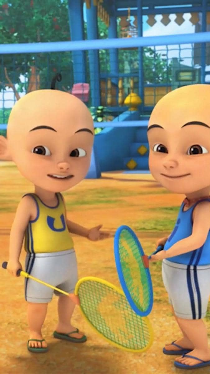 Dua karakter animasi kembar sedang bermain badminton di halaman rumah dengan raket berwarna kuning dan biru.