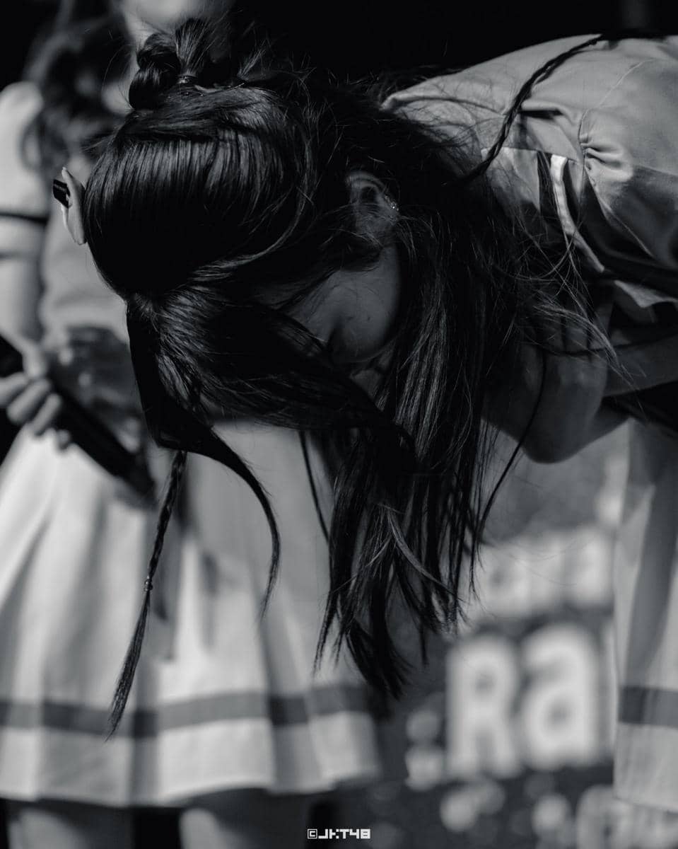Seorang anggota grup idola perempuan membungkuk di atas panggung dengan rambut terurai dan mengenakan kostum pertunjukan.