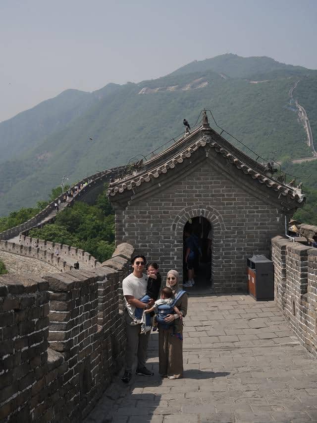 Sebuah keluarga berpose di Tembok Besar Cina dengan latar pegunungan hijau dan bangunan batu berarsitektur tradisional.