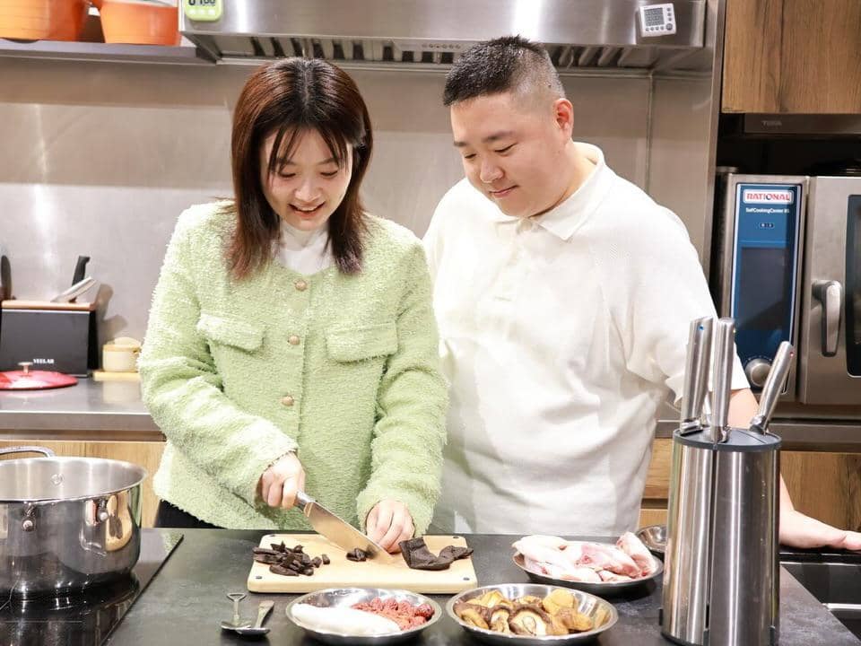Seorang wanita memotong bahan makanan di dapur modern sambil tersenyum, ditemani seorang pria yang berdiri di sampingnya.