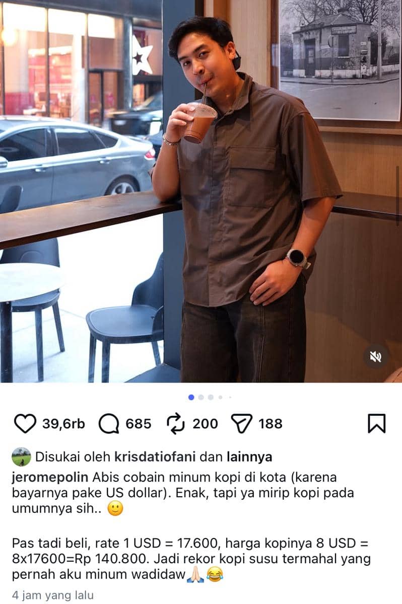 Seorang pria mengenakan kemeja abu-abu sedang berdiri di dekat jendela kafe sambil meminum kopi susu dengan sedotan plastik.