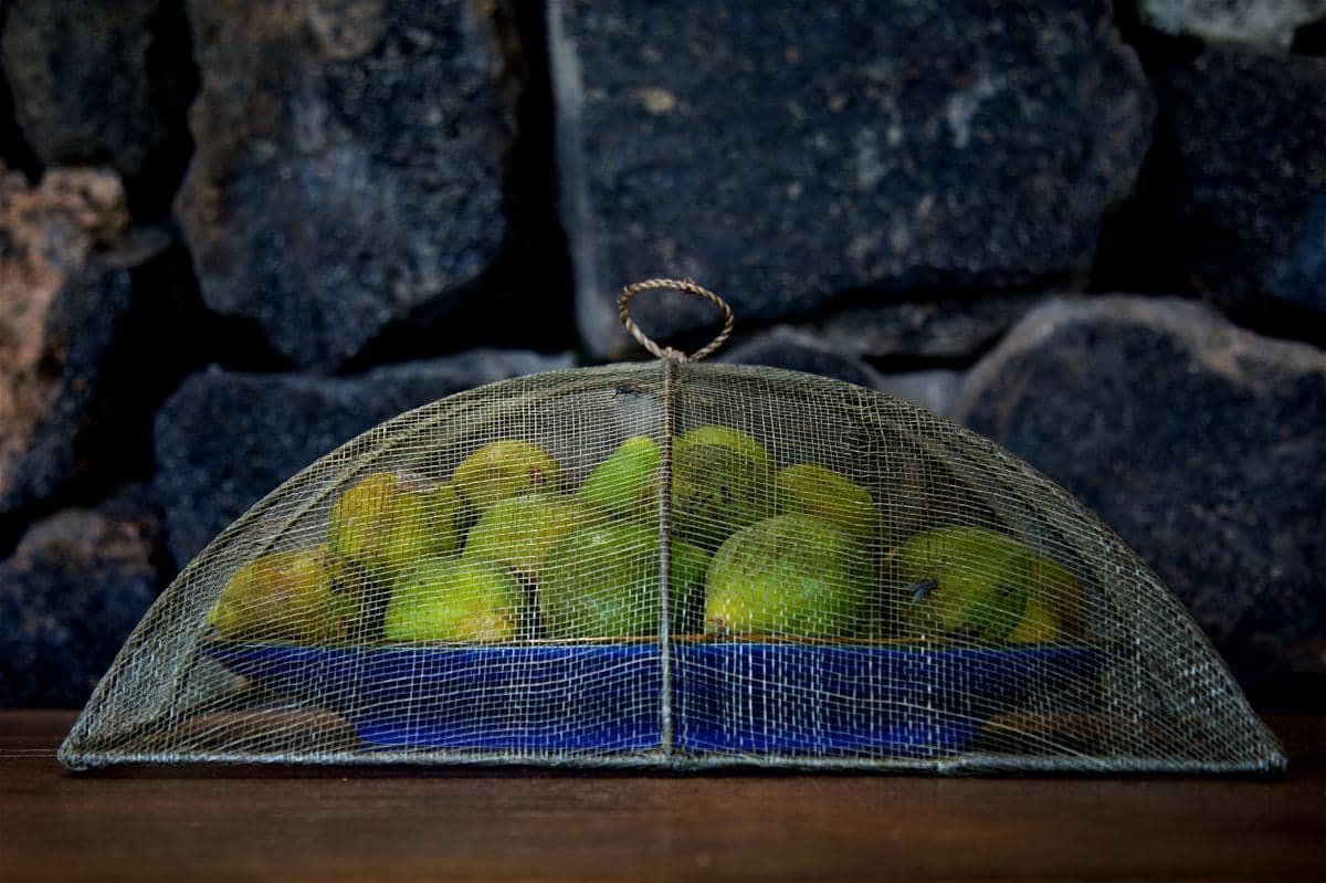Tudung saji berbahan jaring menutupi piring berisi buah pir hijau di atas meja kayu dengan latar dinding batu gelap.