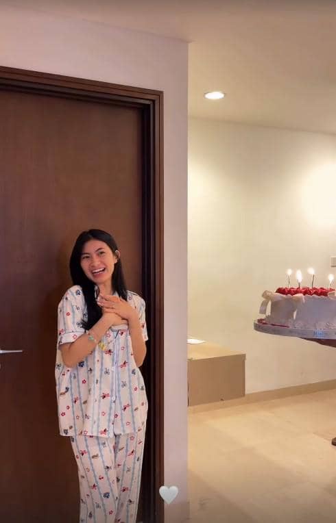 Seorang wanita tersenyum bahagia mengenakan piyama bermotif berdiri di dekat pintu dengan kue ulang tahun berlampu lilin di meja.