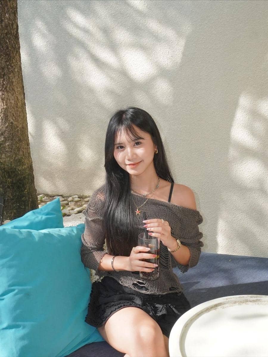 Seorang perempuan berambut panjang duduk santai di luar ruangan sambil memegang minuman di gelas, dengan latar dinding terang dan bantal biru.
