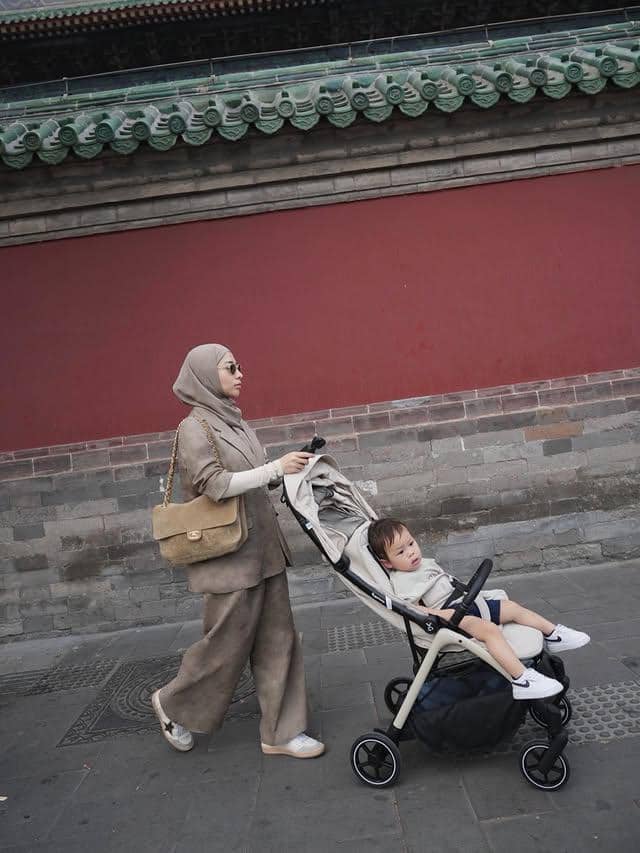 Seorang wanita berhijab mendorong stroller berisi anak kecil di depan dinding merah bergaya arsitektur tradisional Tiongkok.