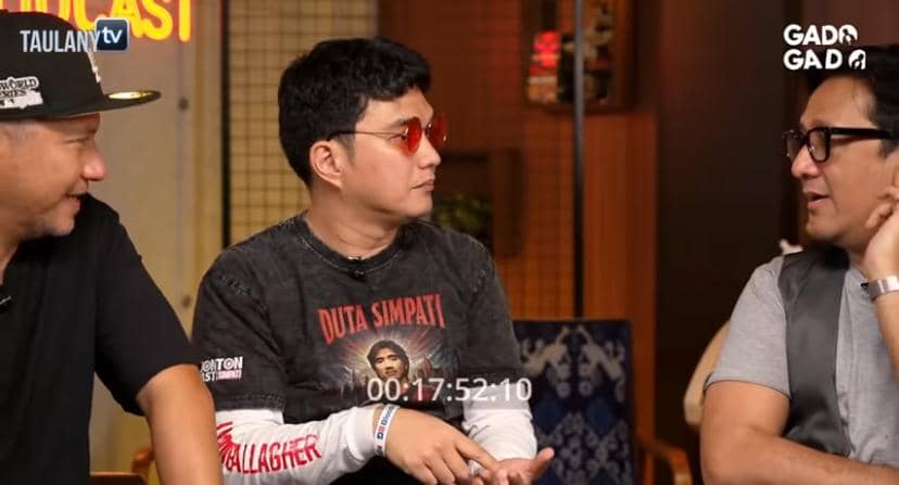 Tiga pria sedang berbincang santai di sebuah studio dengan latar bertuliskan Taulany TV dan Gado Gado, salah satunya mengenakan kacamata merah.