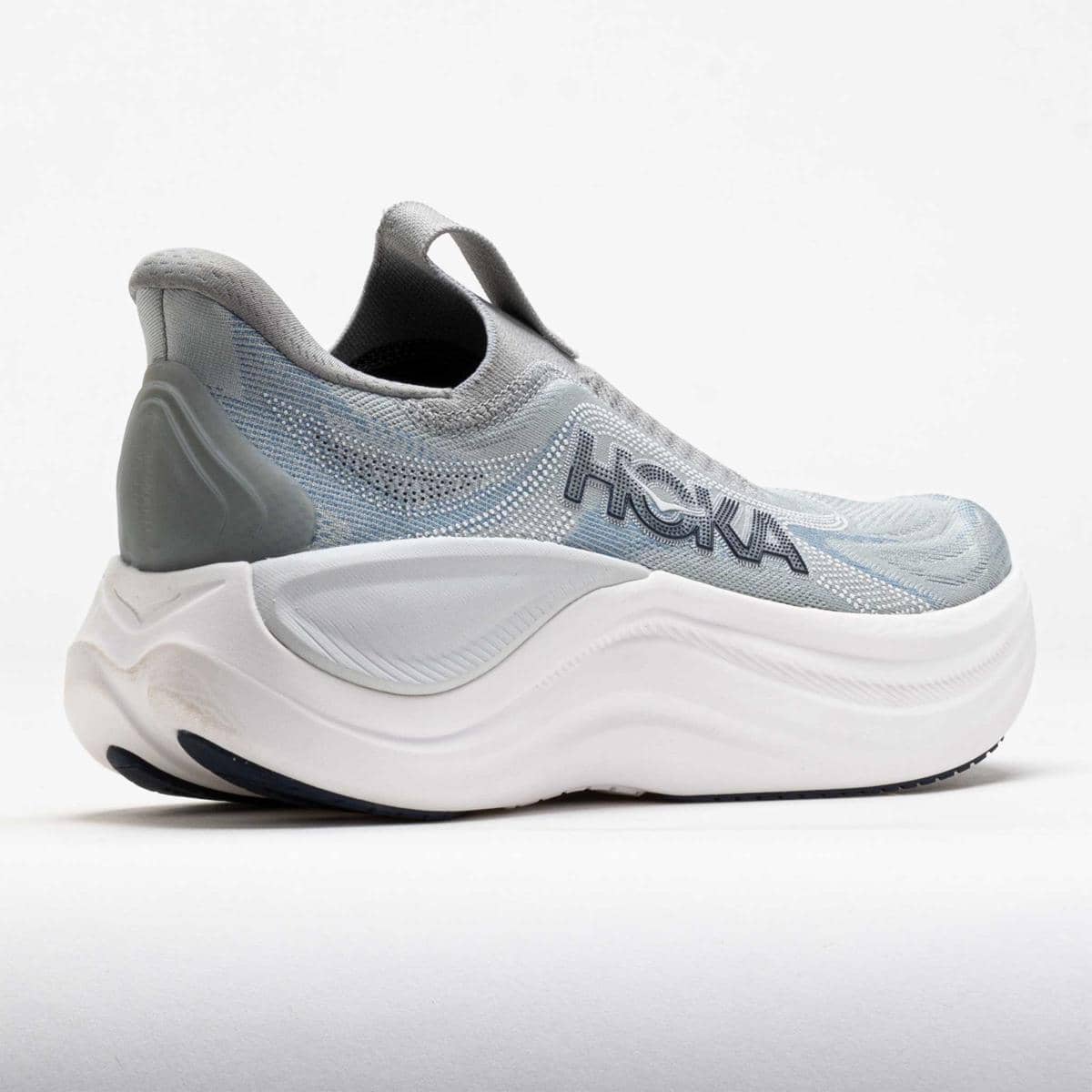 Sepatu Hoka Skyward Laceless berwarna abu-abu dengan desain tanpa tali dan sol tebal berwarna putih untuk kenyamanan maksimal.