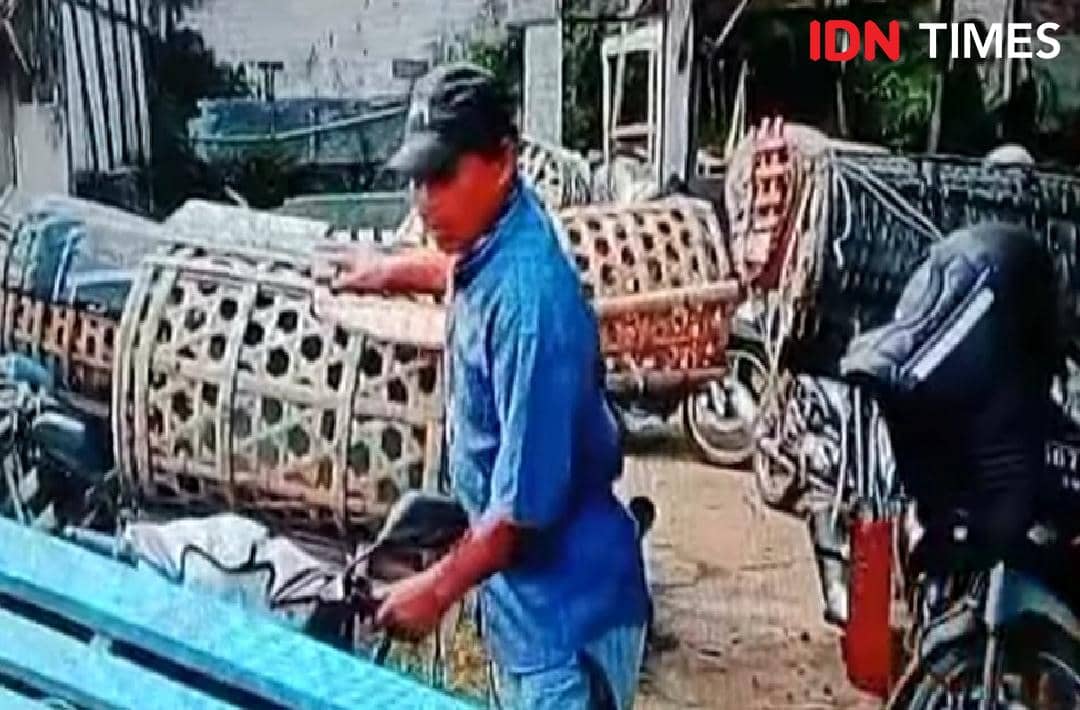 Seorang pria mengenakan kemeja biru dan topi hitam sedang berada di area parkir pasar hewan Ajibarang dengan deretan motor berkeranjang bambu.