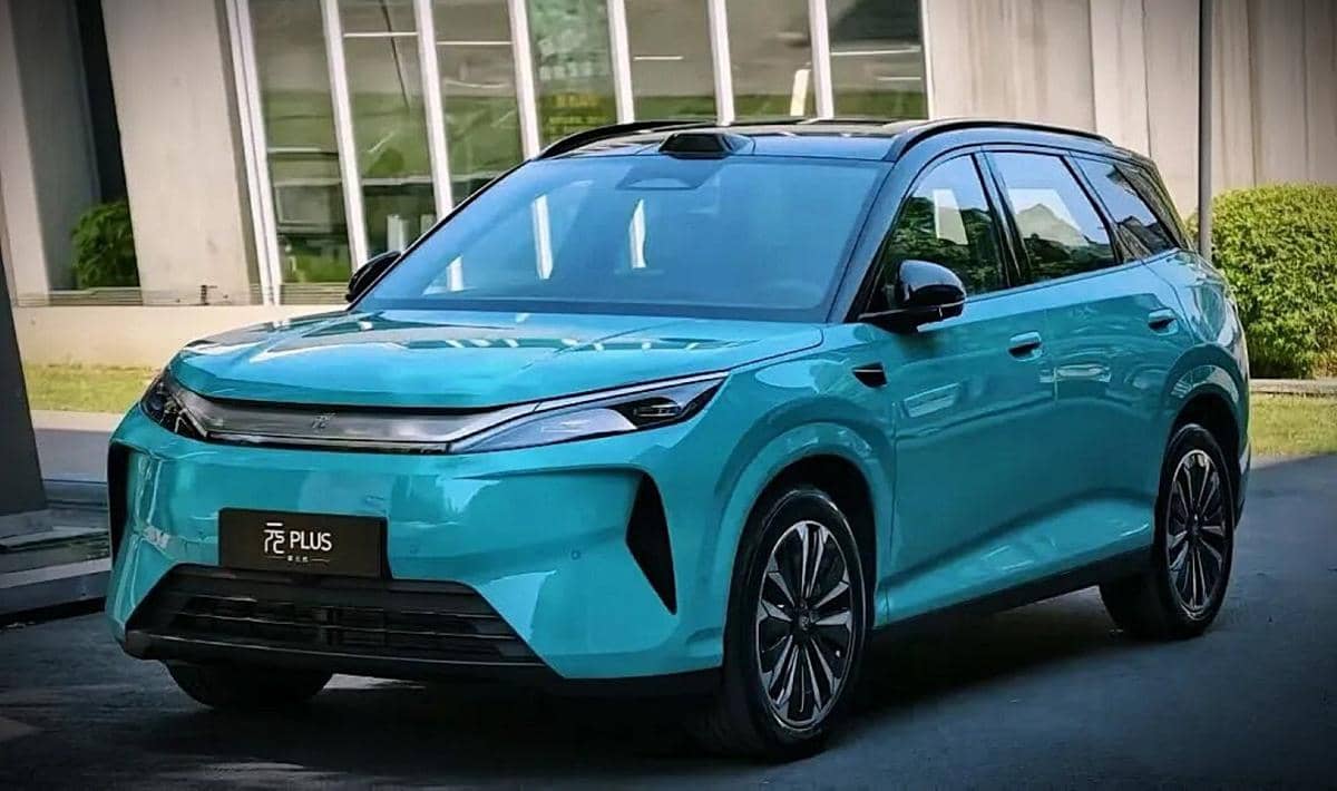 Mobil SUV listrik berwarna biru toska dengan desain modern dan lampu depan ramping diparkir di depan gedung kaca.
