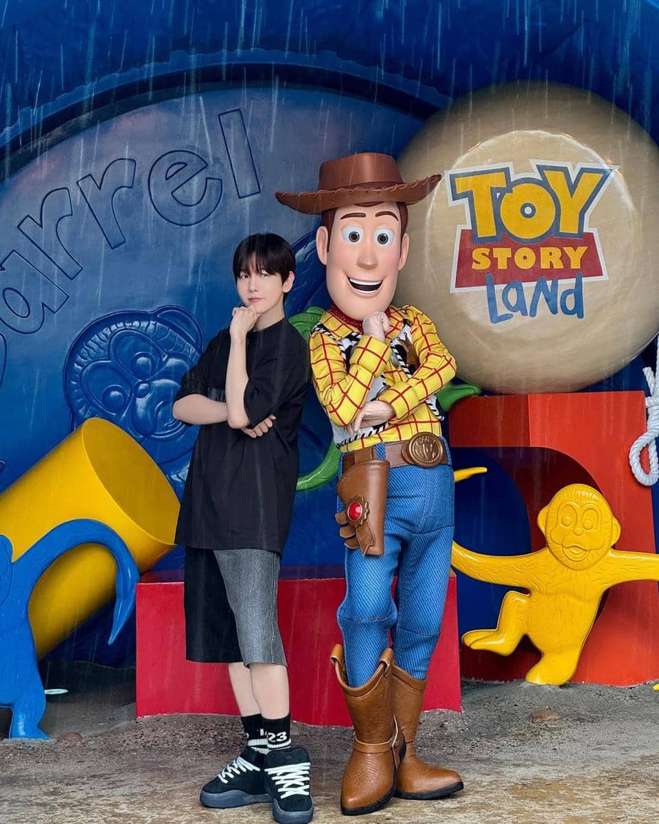 Seseorang berpose di samping karakter Woody dari Toy Story di area Toy Story Land, mengenakan outfit kasual hitam dan sepatu sneakers.