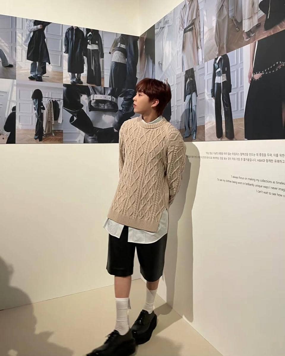 Seorang pria mengenakan sweater rajut krem, kemeja putih, celana pendek hitam, dan sepatu hitam berdiri di galeri dengan dinding bergambar fashion.