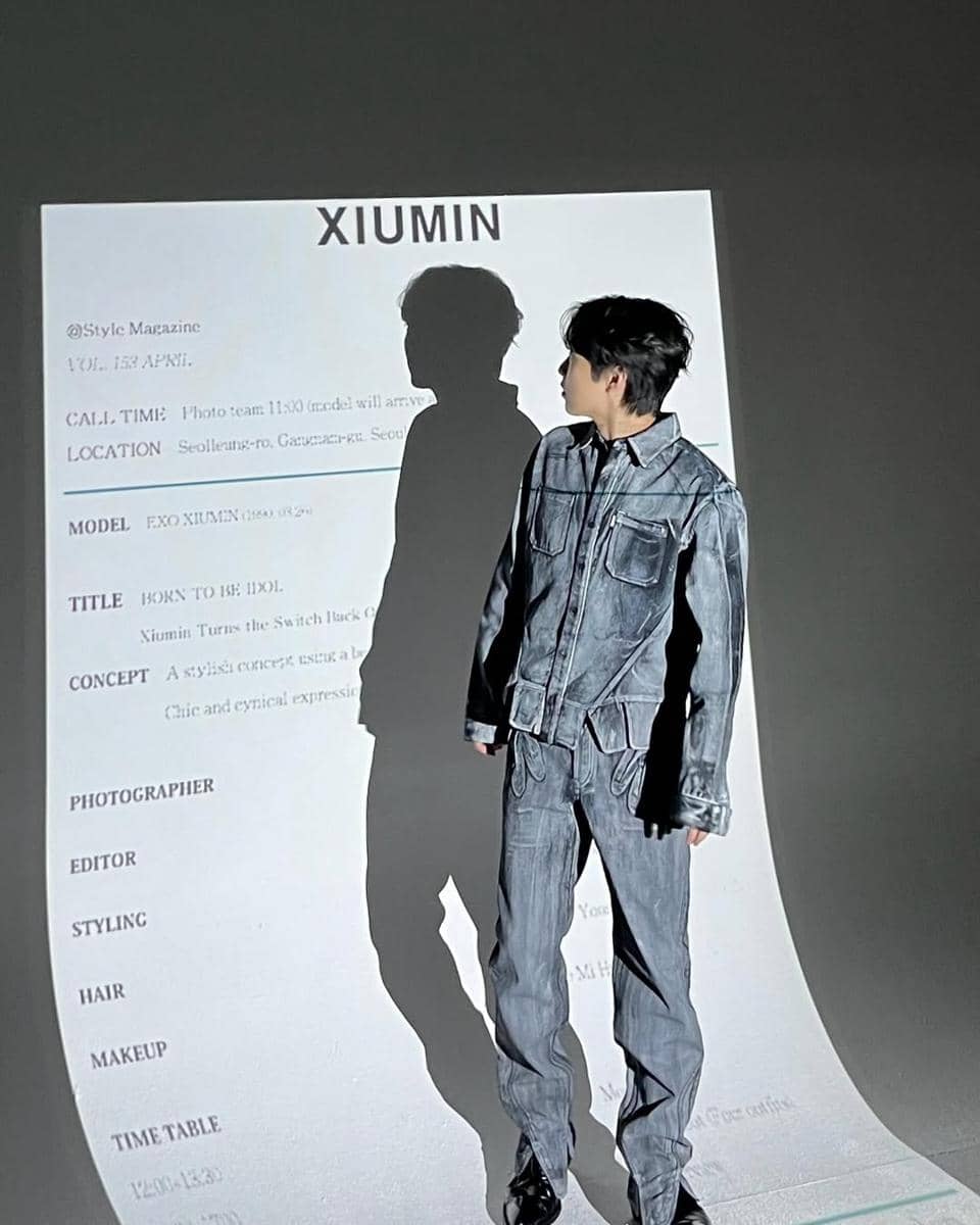 Seorang pria mengenakan setelan denim abu-abu berdiri di depan proyeksi bertuliskan 'XIUMIN' dengan bayangan dirinya di dinding.