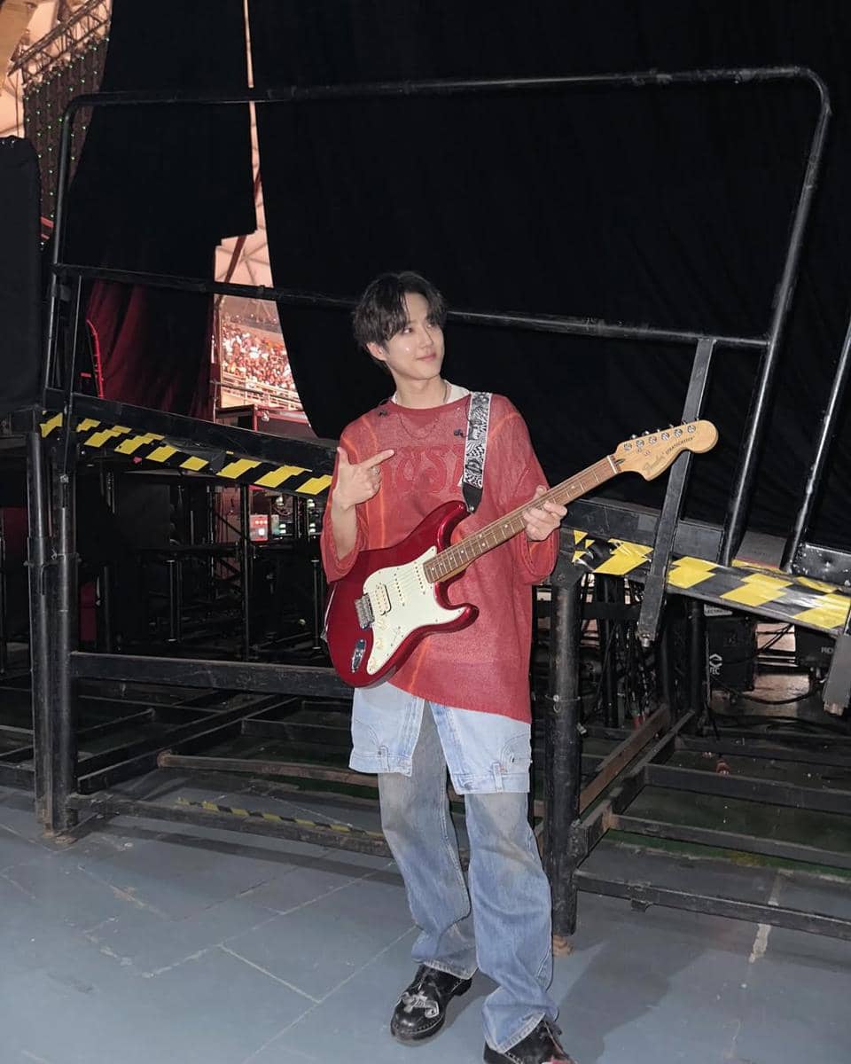 Seorang pria mengenakan kaus merah longgar dan celana jeans longgar sambil memegang gitar listrik merah di belakang panggung konser.