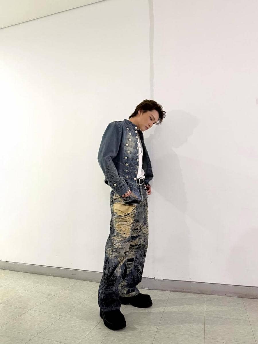 Seorang pria mengenakan jaket denim biru dengan detail kancing perak dan celana denim motif abstrak berdiri di ruangan berwarna putih.