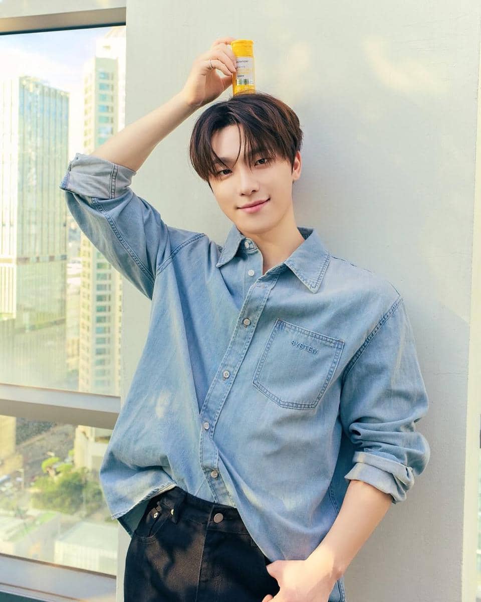 Seorang pria mengenakan kemeja denim biru muda dan celana hitam berdiri di balkon gedung sambil memegang botol kecil di atas kepalanya.