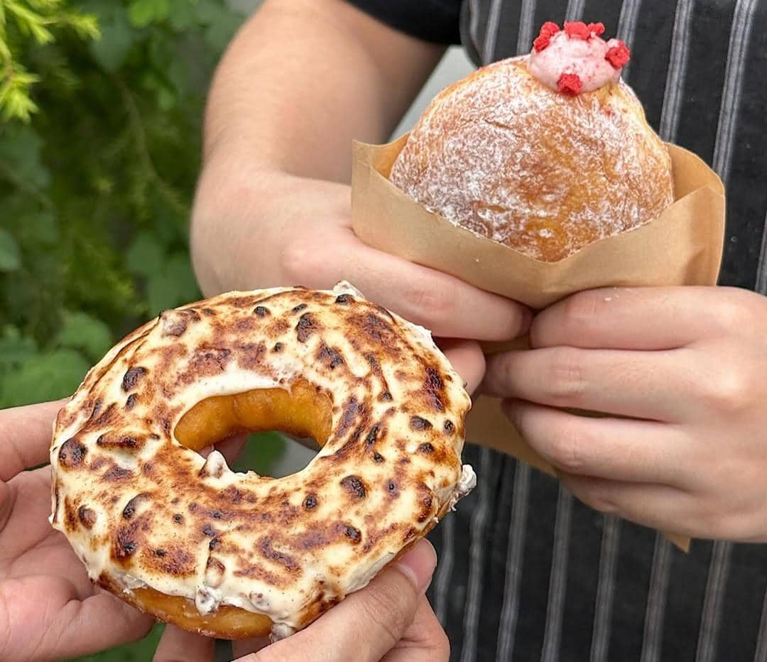 Dua donat artisan, satu dengan topping krim panggang dan satu donat isi krim stroberi, dipegang oleh dua orang.