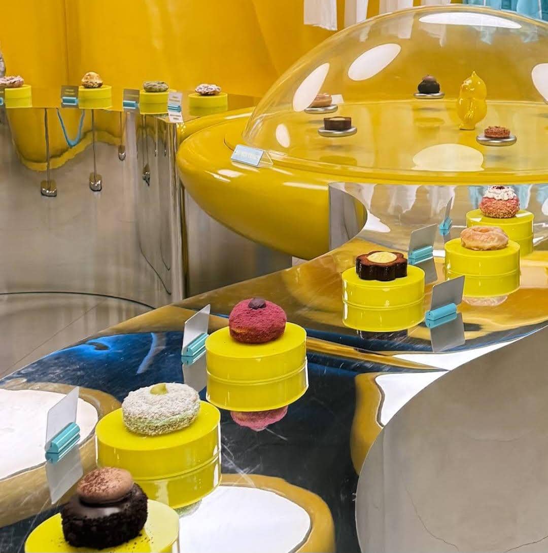 Deretan kue dan pastry berwarna-warni dipajang di atas meja logam reflektif dengan desain futuristik bernuansa kuning cerah.