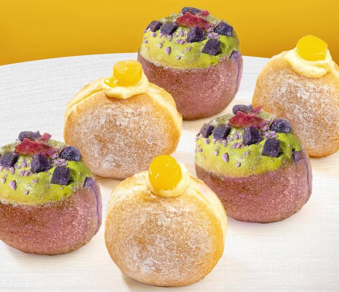 Enam donat lembut dengan topping krim hijau, potongan ubi ungu, dan krim kuning di atas meja berwarna terang berlatar kuning.