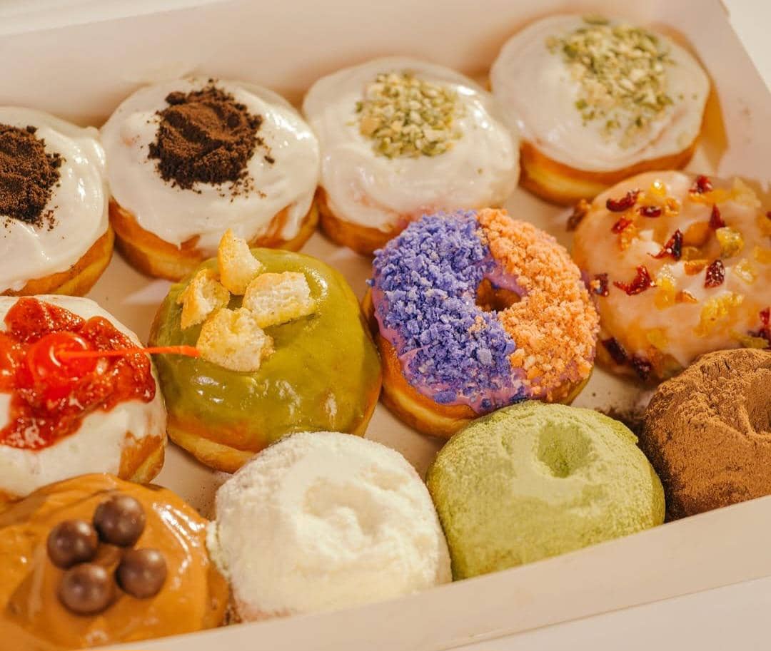 Beragam donat warna-warni dengan berbagai topping seperti krim, cokelat, matcha, dan buah kering tersusun dalam kotak putih.