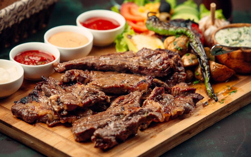 Potongan daging sapi bulgogi panggang disajikan di atas talenan kayu dengan saus, sayuran segar, dan cabai panggang di sekitarnya.