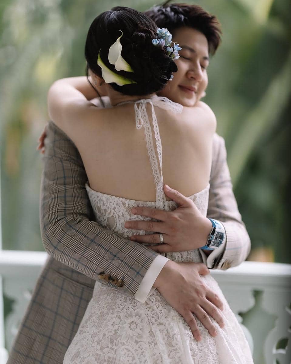 Pasangan pengantin berpelukan dengan penuh kasih, pengantin wanita mengenakan gaun renda putih dan hiasan bunga di rambutnya.