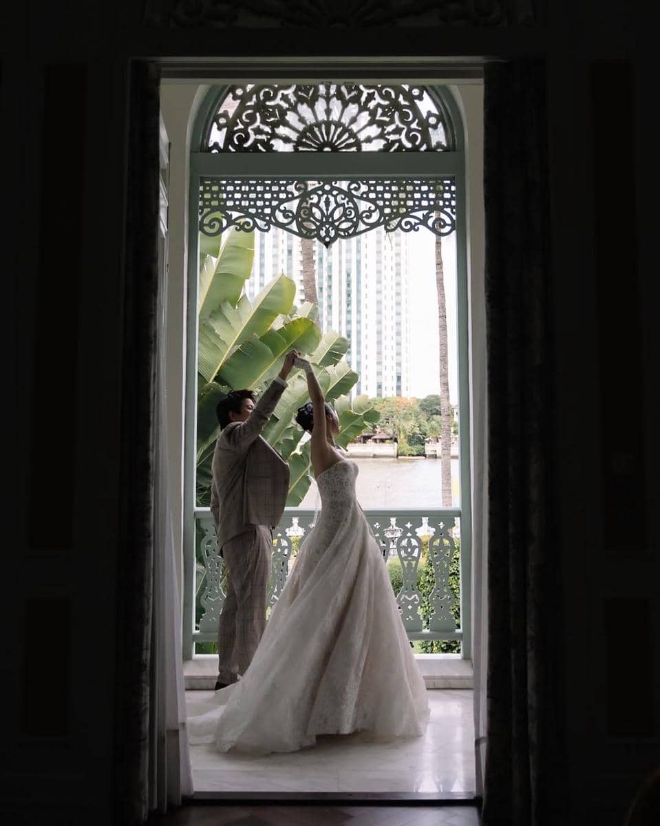 Sepasang pengantin menari di balkon dengan latar pepohonan tropis dan gedung tinggi, mengenakan gaun dan setelan jas elegan.