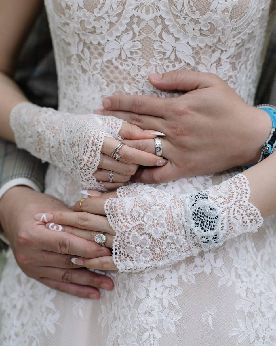 Tangan pasangan pengantin saling berpegangan dengan cincin pernikahan, mengenakan gaun renda putih yang elegan.
