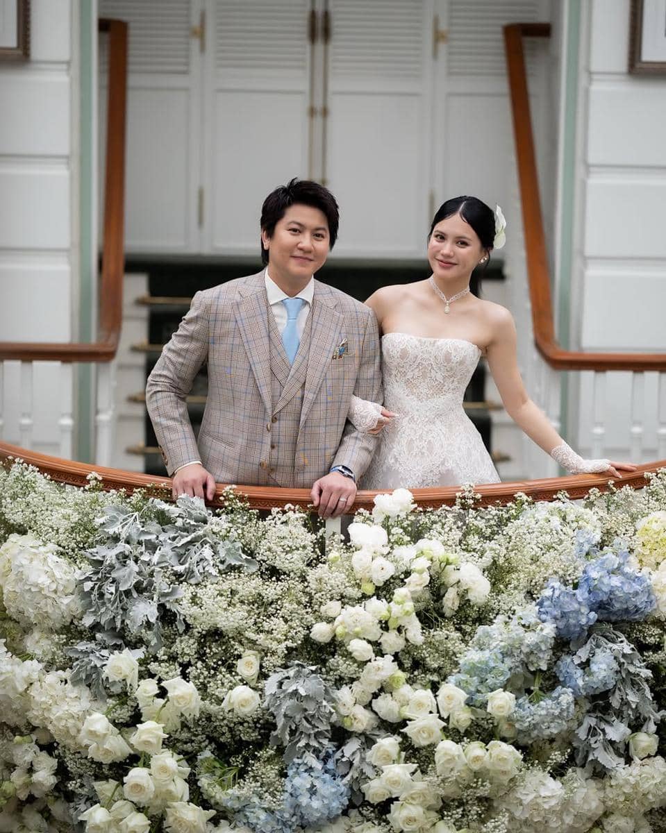 Sepasang pengantin berpose di tangga berhias bunga putih dan biru, sang mempelai wanita mengenakan gaun renda tanpa tali dan mempelai pria bersetelan jas abu-abu bermotif kotak.