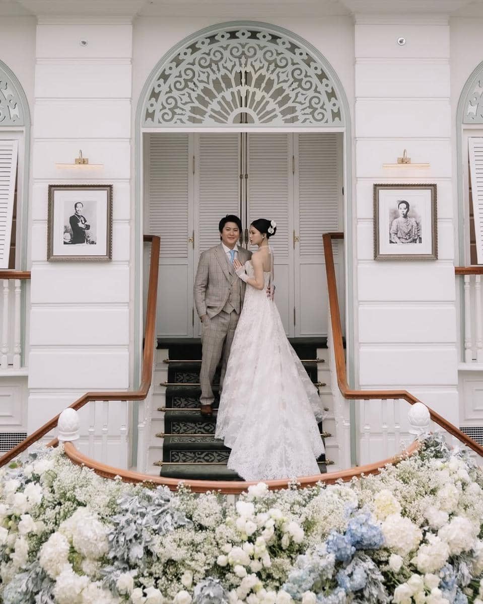 Pasangan pengantin berpose di tangga gedung bergaya klasik dengan dekorasi bunga putih dan biru yang elegan di sekitarnya.
