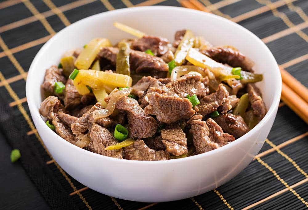 Semangkuk daging sapi lada hitam dengan potongan paprika dan bawang disajikan di mangkuk putih di atas alas bambu hitam.