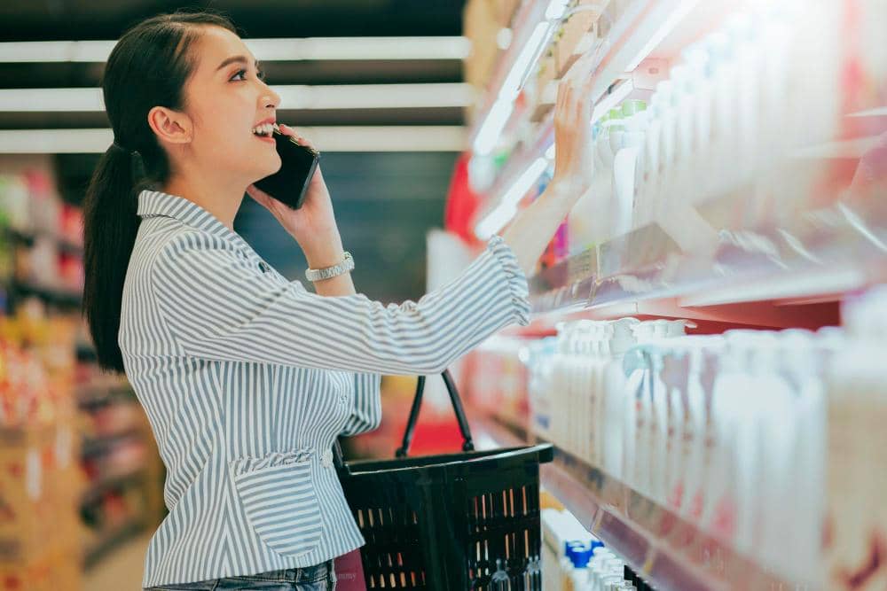Perempuan tersenyum sambil berbicara di telepon dan memilih produk di rak supermarket dengan membawa keranjang belanja hitam.