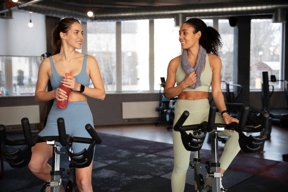 Dua wanita berolahraga bersama di gym, mengenakan pakaian olahraga dan berbincang sambil menggunakan sepeda statis.