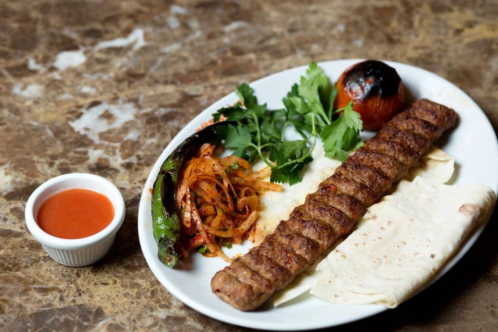 Sepiring kebab daging panggang disajikan dengan roti pipih, bawang berbumbu, cabai panggang, tomat bakar, dan saus merah.