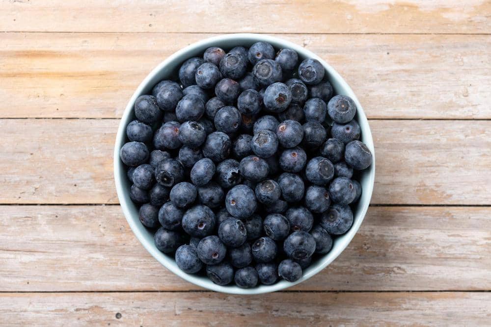 Ilustrasi buah blueberry segar berwarna biru tua dengan lapisan putih alami, tersusun di antara daun hijau.