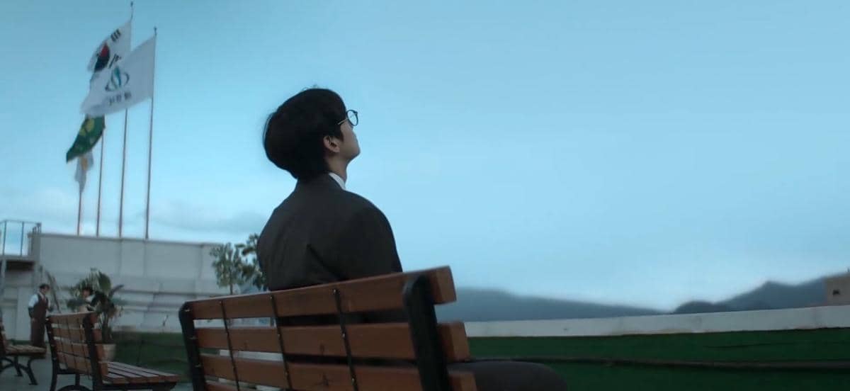 Seorang pria duduk di bangku taman menghadap langit biru dengan latar bendera Korea Selatan dan pegunungan di kejauhan.