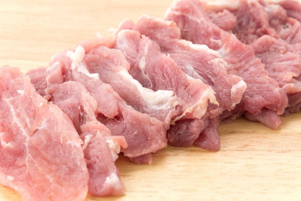 Beberapa potong daging mentah berwarna merah muda dengan serat lemak putih tersusun di atas talenan kayu terang.