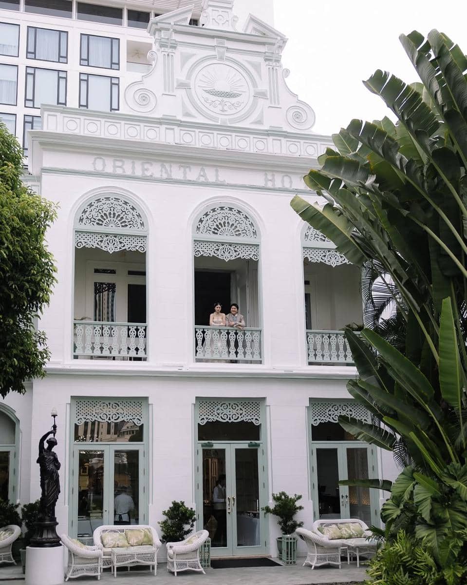 Pasangan berdiri di balkon gedung bergaya kolonial berwarna putih dengan ornamen klasik dan tanaman tropis di sekitarnya.