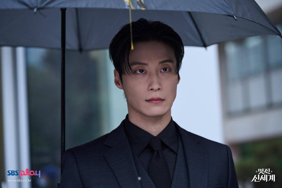 Seorang pria mengenakan setelan jas hitam memegang payung di bawah hujan dalam cuplikan drama Korea My Royal Nemesis.