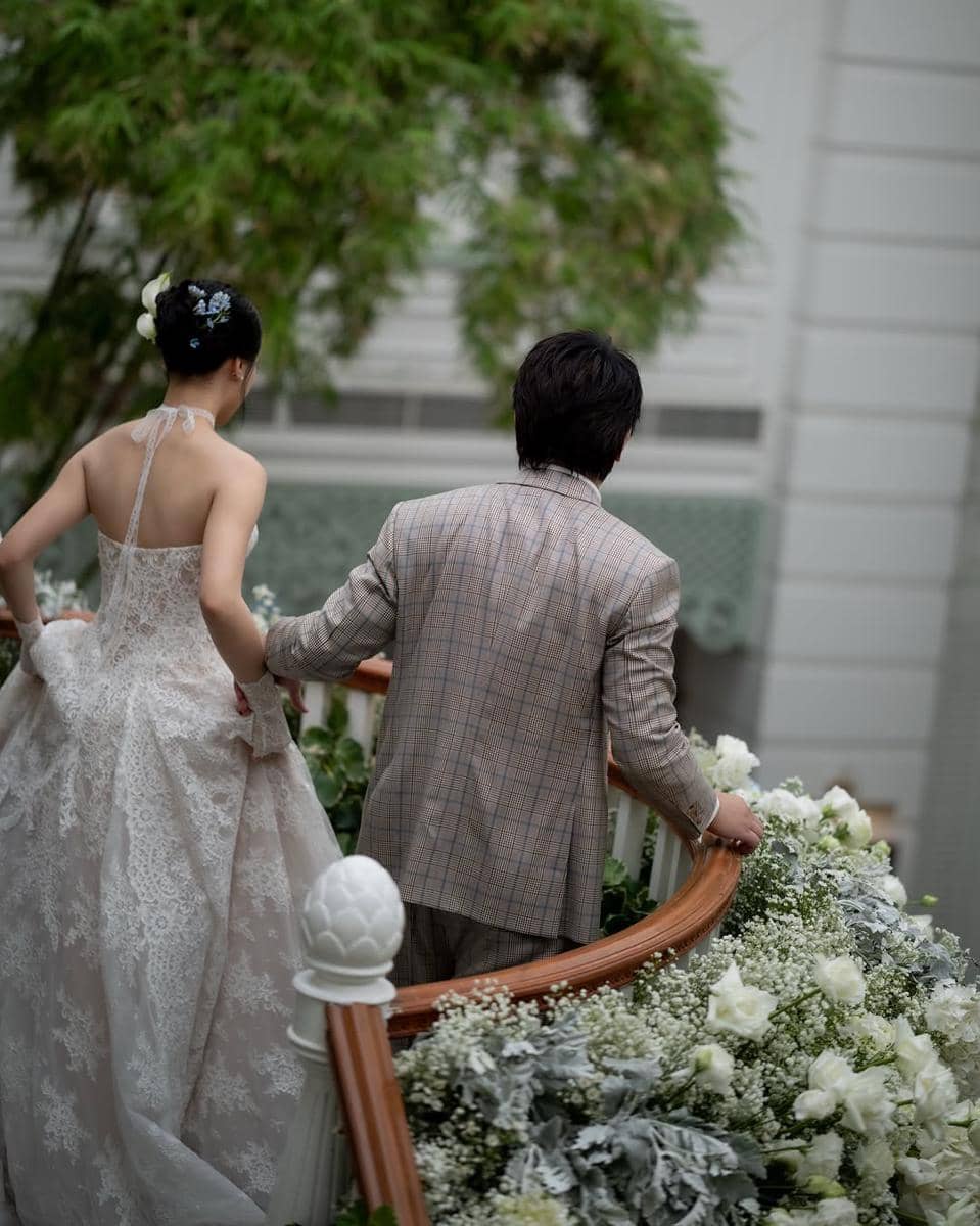 Sepasang pengantin berjalan menuruni tangga berhias bunga putih, sang mempelai wanita mengenakan gaun renda elegan dan pria bersetelan jas kotak-kotak.
