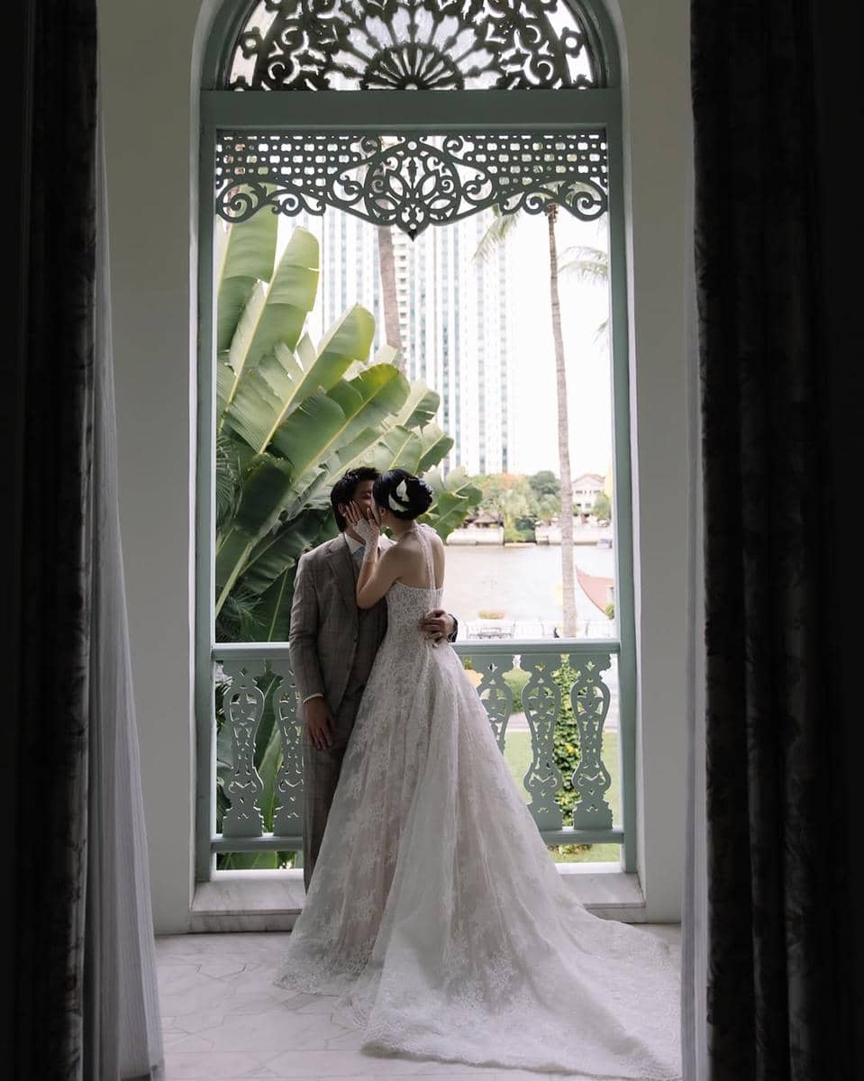 Pasangan pengantin berpose romantis di balkon berhias ukiran, sang mempelai wanita mengenakan gaun putih panjang dengan latar gedung tinggi.