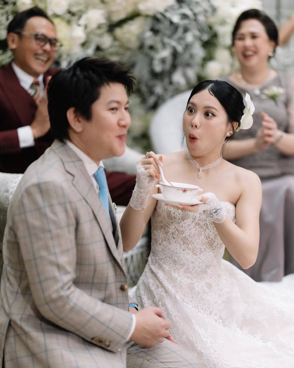 Pasangan pengantin tersenyum bahagia saat upacara pernikahan, sang mempelai wanita mengenakan gaun renda putih dan memegang mangkuk.