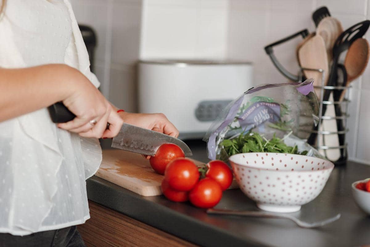 Seseorang memotong tomat segar di dapur dengan pisau di atas talenan, di samping mangkuk dan sayuran hijau untuk membuat salad.