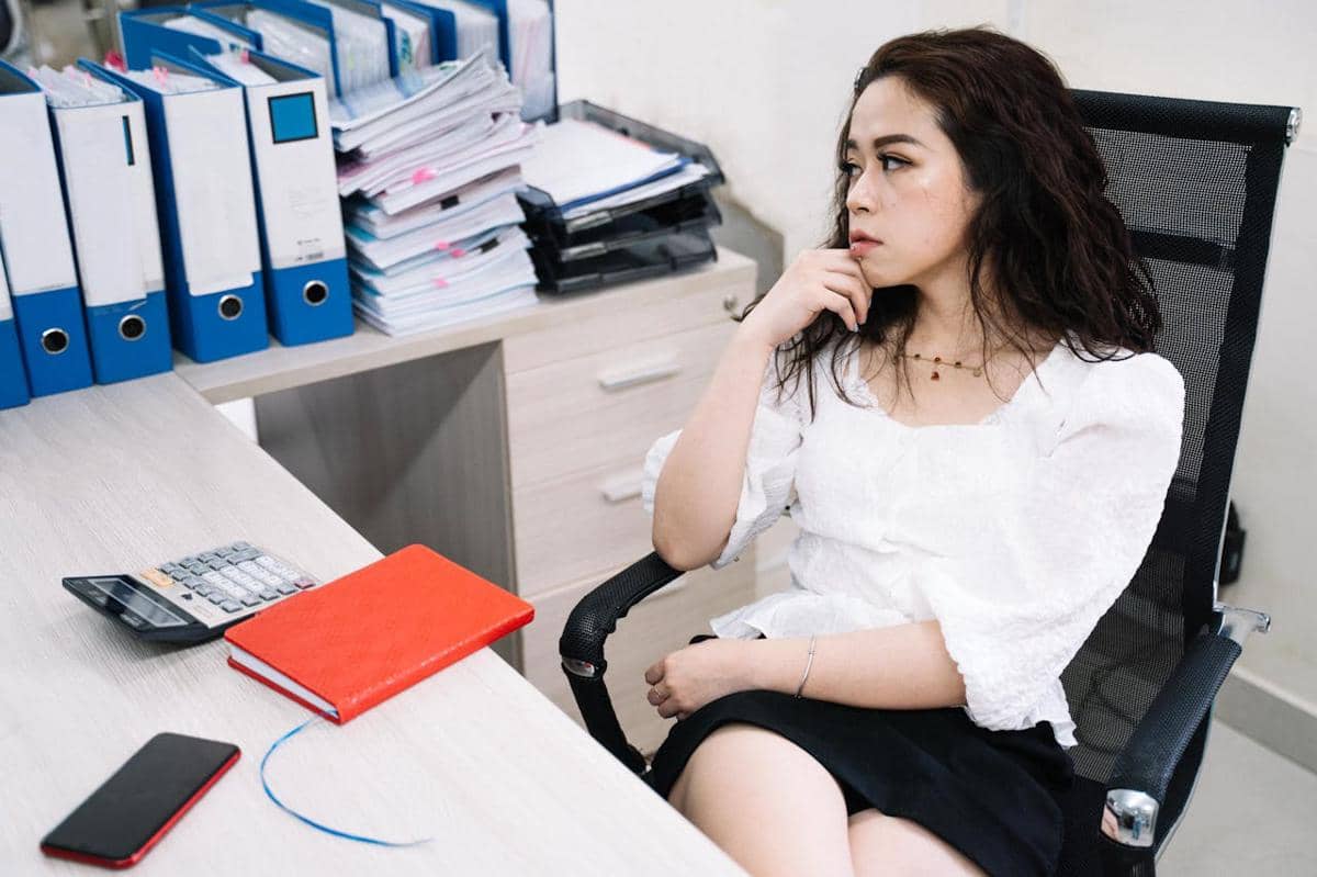 Seorang wanita duduk di kursi kantor dengan ekspresi bosan, dikelilingi tumpukan berkas, buku catatan merah, dan kalkulator di meja.