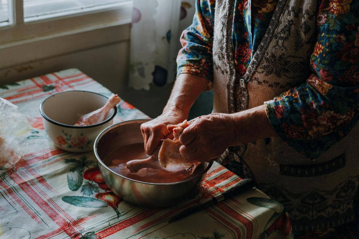 Seseorang mencuci potongan daging ayam mentah di dalam mangkuk logam di atas meja dapur dengan taplak bermotif warna-warni.