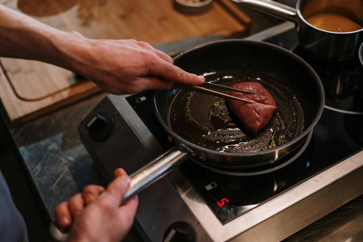 Seseorang sedang memanggang potongan daging di atas wajan berisi minyak panas di kompor dapur modern.