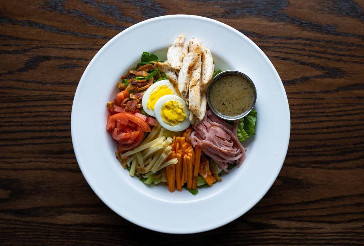 Salad Cobb ala Yunani dengan potongan ayam, telur rebus, ham, keju, tomat, wortel, dan saus dressing di atas piring putih.